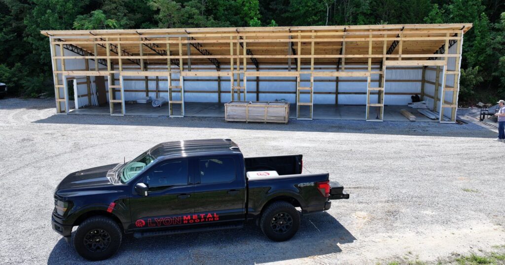 A large post-frame pole barn under construction featuring engineered wood trusses, metal roofing and siding, treated lumber framing, and a Lyon Metal Roofing truck parked in front. This custom pole barn building showcases quality pole barn materials for agricultural, commercial, and residential construction projects.